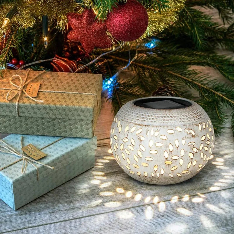A round, off-white outdoor lantern with decorative oval cutouts emits a warm white glow, casting star-shaped shadows on a light wooden surface. The lantern has a dark, flat top, likely a solar panel. It sits near wrapped gifts and the branches of a decorated Christmas tree with ornaments and lights.