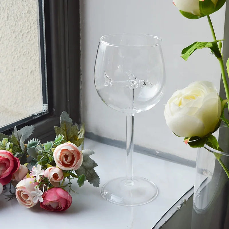 A clear, empty wine glass with a small, suspended glass shark figurine inside. The glass is on a windowsill with flowers nearby.