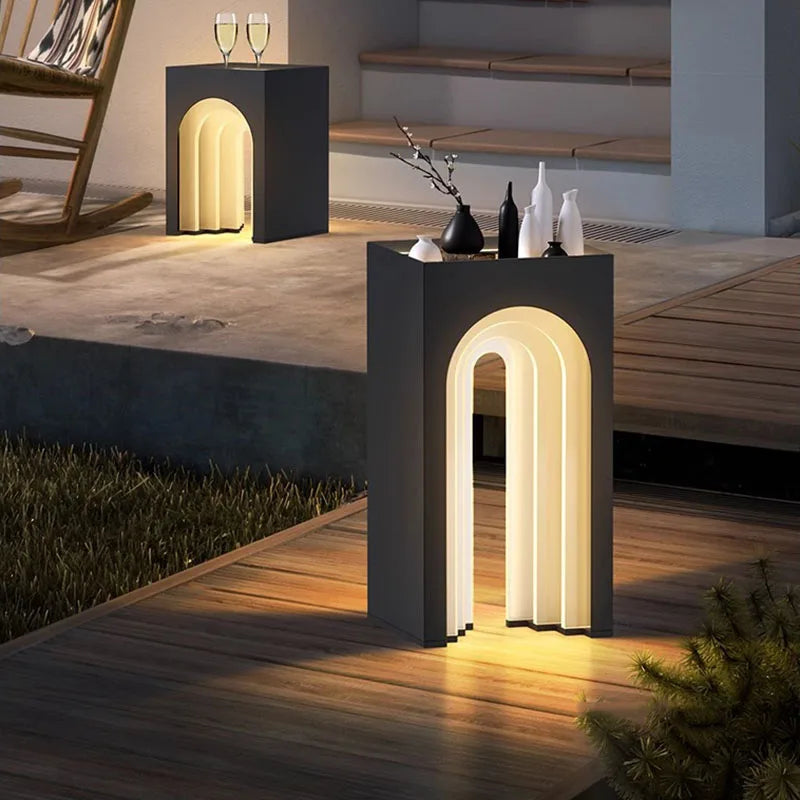 Two modern, dark gray solar-powered archway glow lamps serve as side tables on a patio. One holds a vase and white bottles, the other two wine glasses.