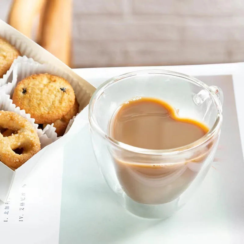 A heart-shaped glass mug of coffee is next to a box of cookies on a white surface.