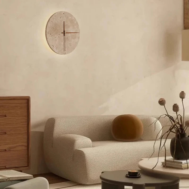 A round wall clock with a light-colored, textured face and simple red hands is mounted on a light beige wall. The clock emits a soft glow from behind. Below, a modern, off-white curved sofa is adorned with a round brown pillow. A dark vase with dried floral stems sits on a round coffee table in front of the sofa, along with a small cup. Part of a wooden cabinet is visible on the left.