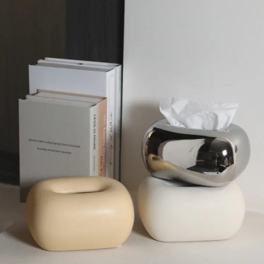A matte yellow, a shiny chrome, and a matte white donut-shaped tissue box holder are stacked next to a stack of books.