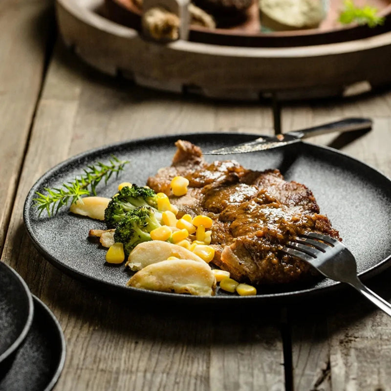A black ceramic plate with a steak, corn, and broccoli, on a rustic wooden table.
