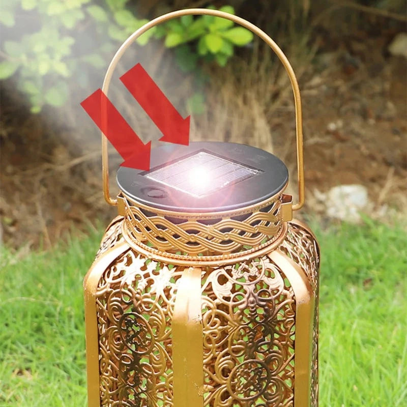 A close-up of a decorative gold-colored metal solar lantern with intricate cutouts shows sunlight shining on its solar panel. Two red arrows point towards the panel. The lantern has a handle and is surrounded by blurred green foliage.