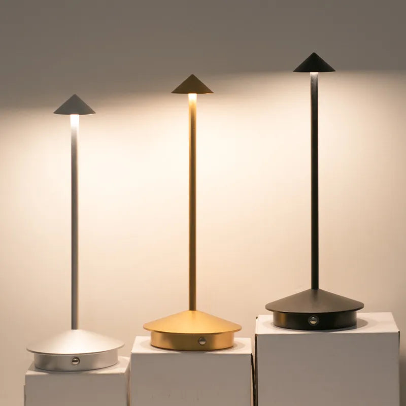 Three cordless table lamps with slender stems and pointed shades stand on white surfaces against a blurred background. The lamps are silver, gold, and black.
