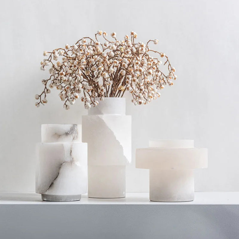 A set of three modern alabaster vases in varying geometric shapes, featuring a smooth white and marbled texture. One of the vases holds a delicate arrangement of dried white berry branches. The vases are displayed on a white surface against a light gray background, creating a minimalist and elegant aesthetic.