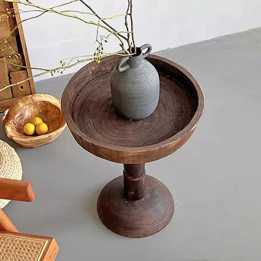 A rustic, dark wood, round side table (Momo Wabi-sabi Log Small Coffee Table) holding a gray vase with bare branches, positioned in a room with a wooden basket containing lemons and a woven stool.