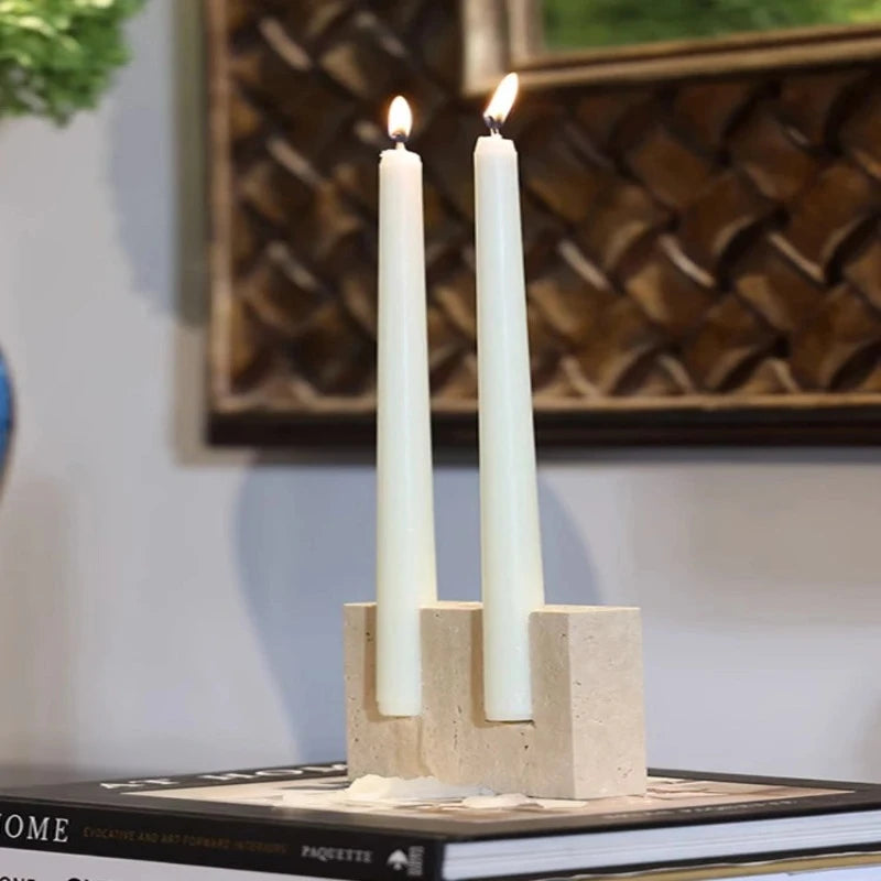 Two lit, off-white taper candles in a textured, light-colored faux travertine stone block holder with cylindrical openings, resting on a book.