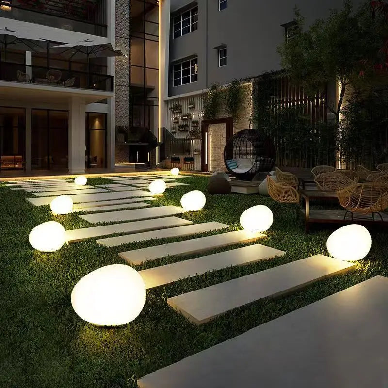 A modern outdoor patio with a stone pathway lined with glowing egg-shaped lights, leading to seating areas and a building.