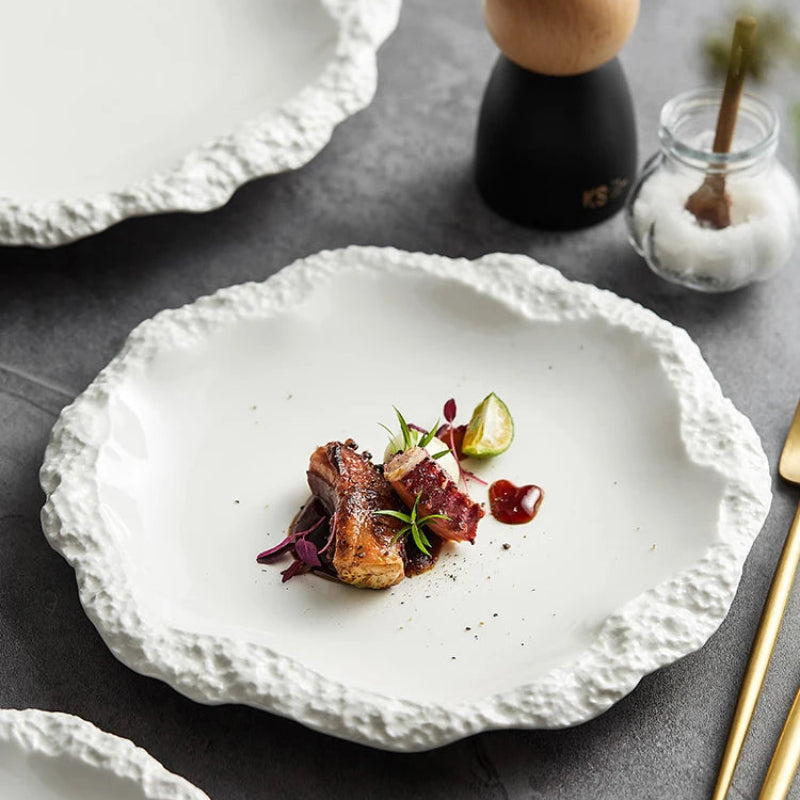 A perfectly seared cut of meat is artfully arranged on a white, textured plate, garnished with a lime wedge and herbs, with a salt shaker and another plate in the background.