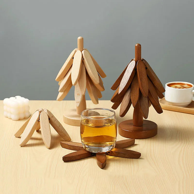 Three wooden trivets are on a light wooden table: two tree-shaped (one light, one dark) and one flat, starburst-shaped with a glass of amber liquid on it. A white candle and a mug are in the background.