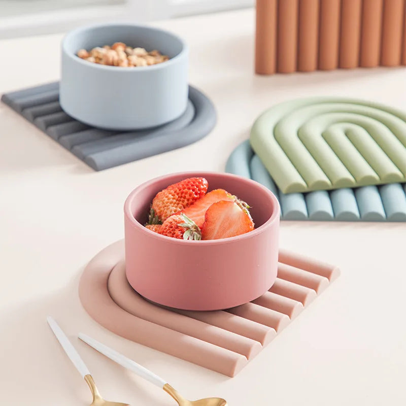 Three colorful silicone trivets with a segmented arch design are arranged on a light-colored surface. A light blue bowl filled with granola sits on a gray trivet. A dusty pink bowl filled with sliced strawberries rests on a matching pink trivet. A muted green trivet lies on a teal blue one. Two white spoons with gold accents are in the foreground. A rust-orange trivet is partially visible in the upper right.