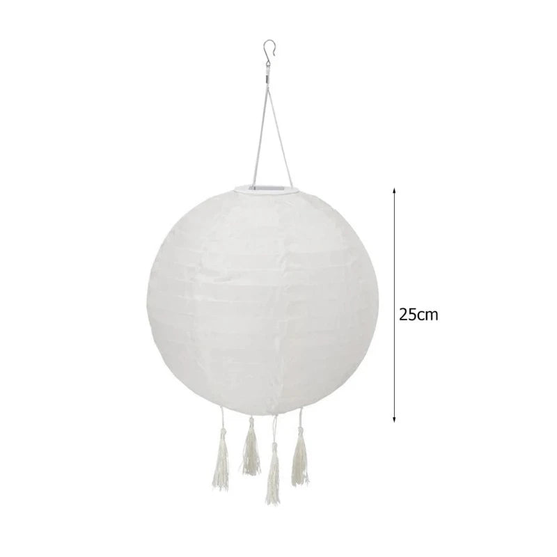 A white, round solar lantern with tassels hangs and is measured at 25cm.