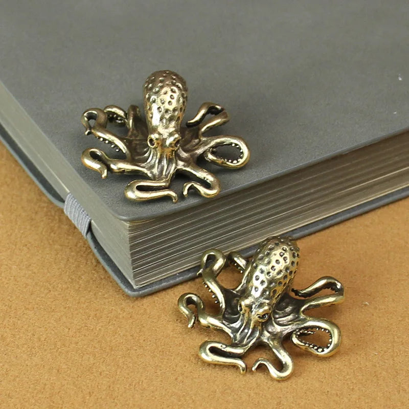 The image shows two small metallic octopus-shaped objects with an antique brass or bronze finish. They are intricately designed with detailed tentacles and textured heads. One of them is placed on top of a closed gray notebook, while the other rests on a brown surface beside it. The objects appear to be decorative, possibly used as bookmarks, charms, or small figurines.