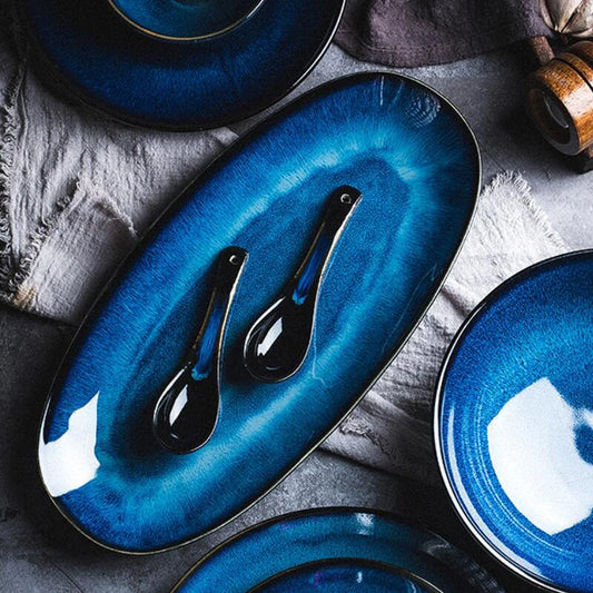 A deep blue oval plate with a swirling white pattern, holding two small, dark-colored spoons. The plate is surrounded by other matching blue plates on a gray, textured fabric.
