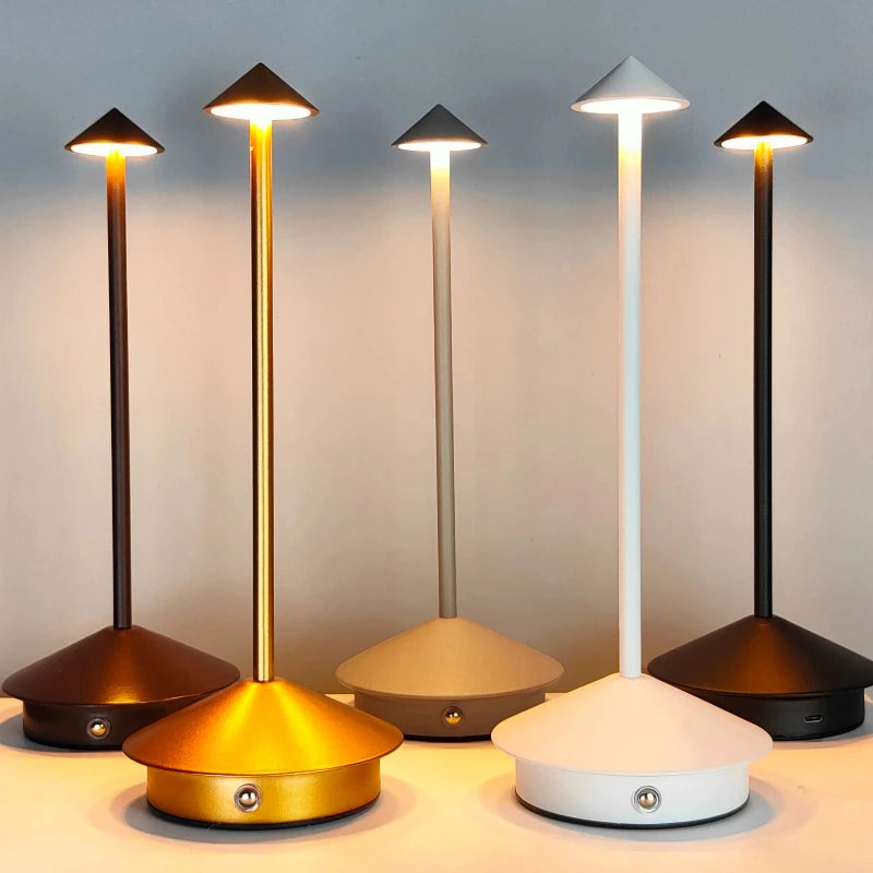 Five cordless table lamps with slender stems and pointed shades in varying metallic and neutral colors are illuminated on a light surface against a muted background. Each lamp emits a different color temperature of light.