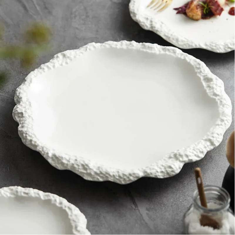 A pristine, white textured plate rests on a dark gray surface, surrounded by other similar plates and a salt shaker.