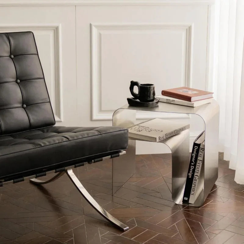A shiny, silver metal side table with rounded corners sits on a dark wood floor next to a black leather chair.
