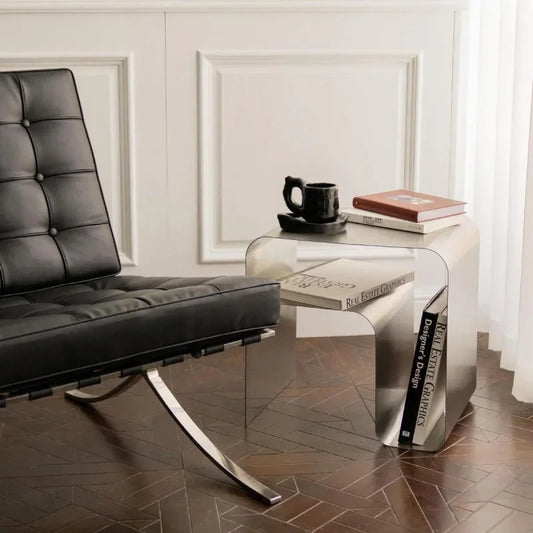 A shiny, silver metal side table with rounded corners sits on a dark wood floor next to a black leather chair.