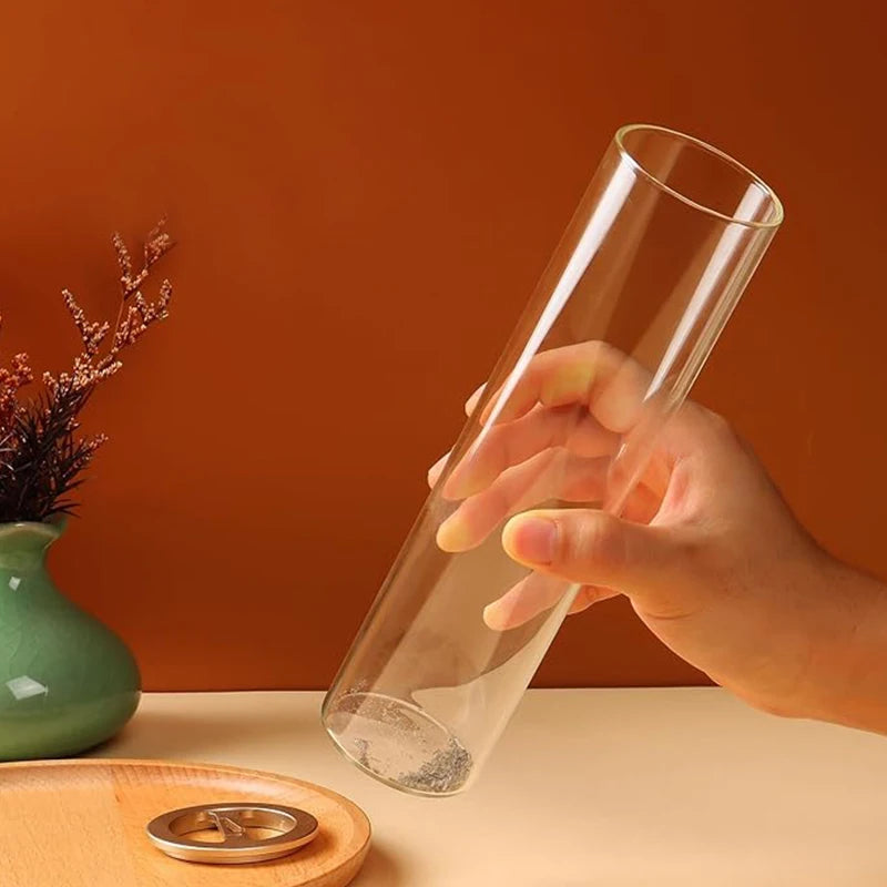 A hand holds a transparent glass cylinder, which appears to be an incense holder. The bottom of the cylinder contains ash, indicating previous use. The metal lid, which likely holds the incense stick, rests on a wooden tray. The background features a warm-toned wall, a green vase with dried plants, and a clean, minimalist setting.