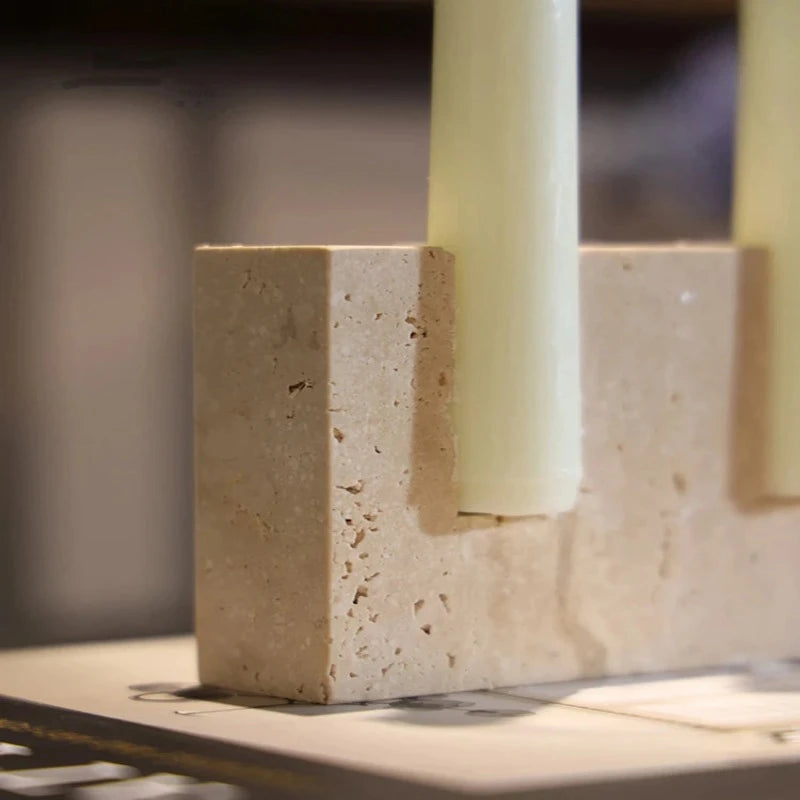A close-up of a textured, light-colored travertine stone block with a cylindrical opening holding a pale yellow taper candle.