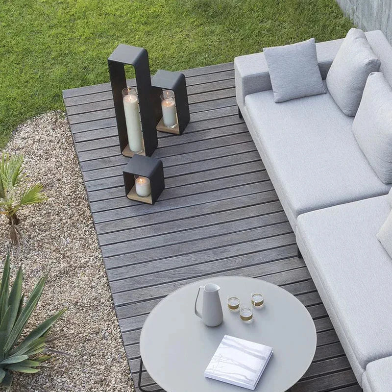 An aerial view of a modern outdoor patio with a gray sectional sofa, a round coffee table with a pitcher and glasses, and three dark, contemporary lanterns with glowing cylindrical lights, arranged on a wooden deck next to a gravel and grass area.