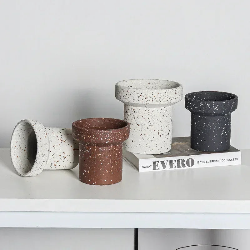 Four small, cylindrical plant pots with slightly flared rims are arranged on a white surface against a light grey background. The pots have different speckled finishes: one is brown with beige speckles, one is white with black speckles, and two are black with white speckles. One of the white speckled pots is lying on its side. Two of the pots rest on a book.