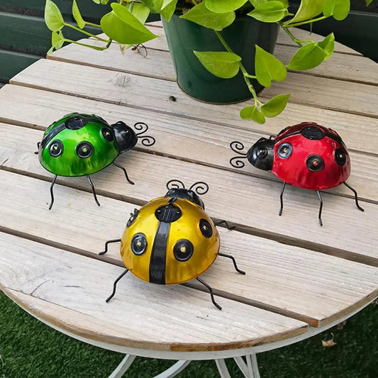 Three decorative metal ladybugs, one green, one red, and one yellow with black stripes, sit on a round wooden table with a potted plant. Each ladybug has black antennae and legs.