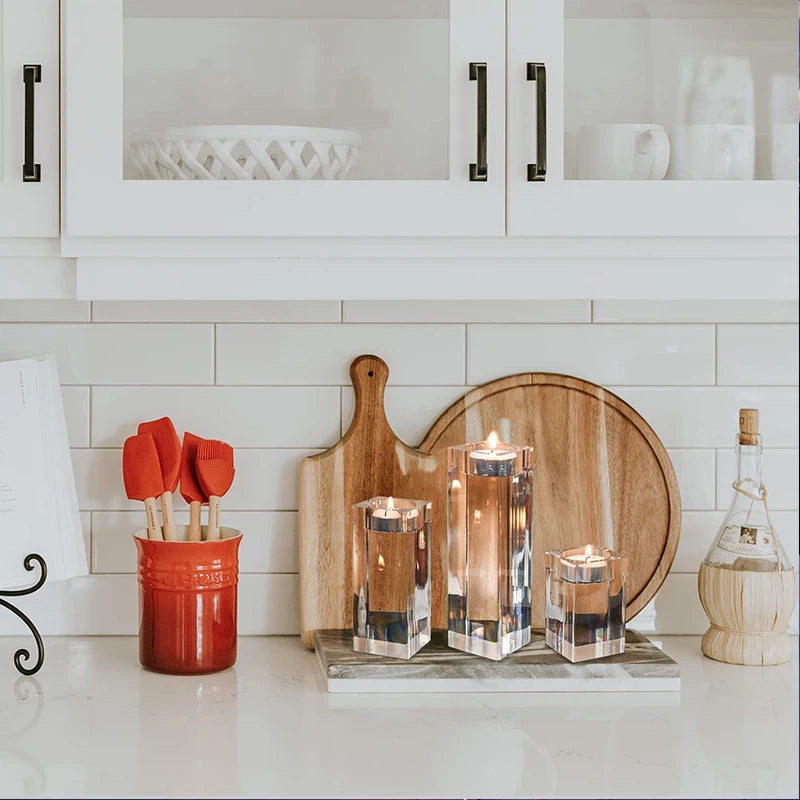 Three clear, multifaceted crystal candle holders of varying heights, each with a lit candle, are arranged on a white countertop in a kitchen setting. They sit on a grey marble cutting board, behind which are two wooden cutting boards. To the left, red silicone kitchen utensils are in a red ceramic holder. White subway tiles form the backsplash, and white cabinets with black handles are above. A clear bottle with a cork stopper is on the right. The candles cast a warm glow.