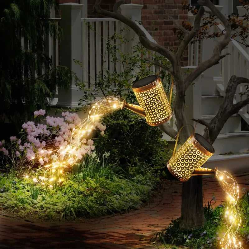 Two decorative, golden watering cans with intricate cutouts are positioned in a garden at dusk or dawn, each illuminated by solar-powered lights within. One watering can is perched on a tree branch, appearing to pour a stream of warm-toned lights onto the greenery below. The other is placed near flowering bushes, with a similar stream of lights extending towards the ground. The illuminated cutouts cast patterns of light, creating a whimsical and enchanting garden scene. A house with white railings is softly