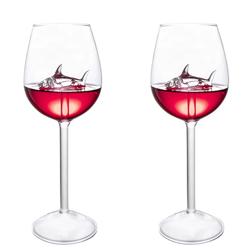 A pair of wine glasses filled with red liquid, each containing a small, clear glass shark figurine.