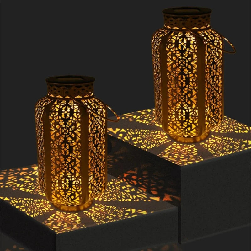 Two decorative metal solar lanterns with intricate cutouts emit a warm yellow light, casting patterned shadows on dark gray surfaces at night. Each lantern has a handle.