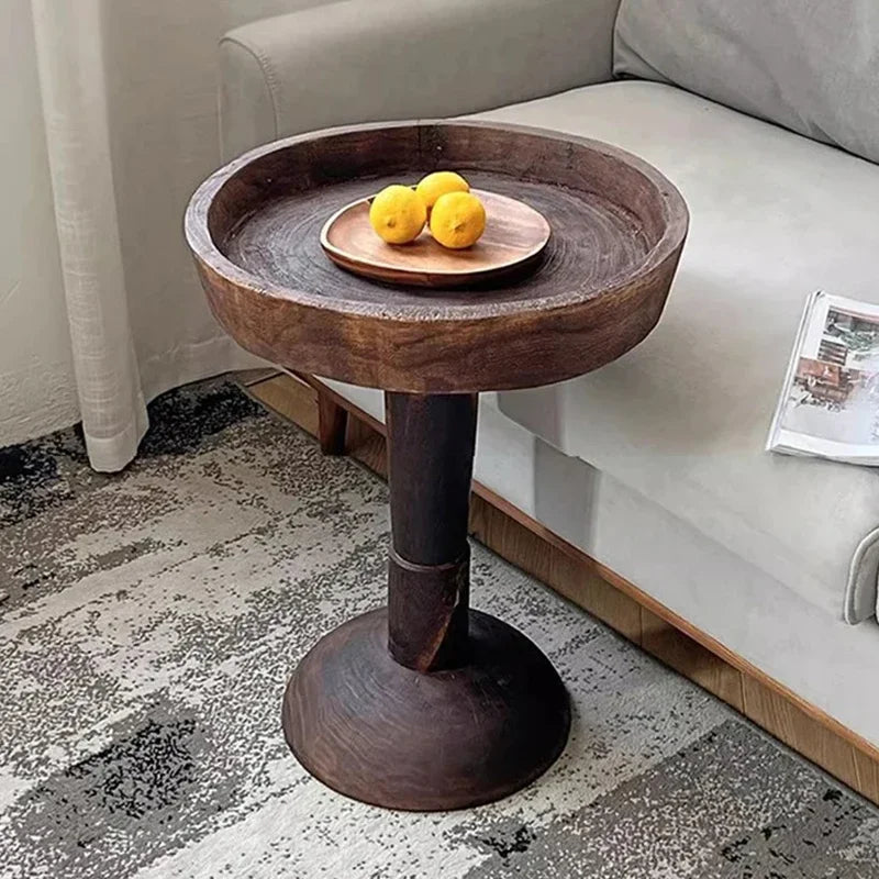 A rustic, dark wood, round side table with a shallow tray top, holding a small plate with three yellow lemons, positioned next to a light-colored sofa on a patterned rug.