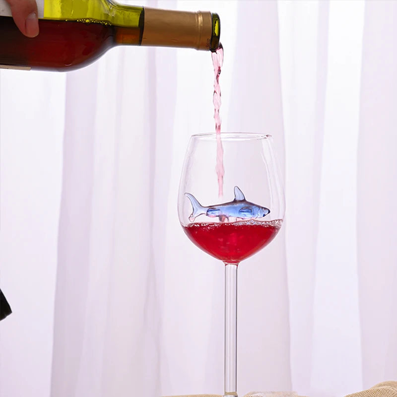 A red liquid being poured from a wine bottle into a wine glass with a small, blue glass shark figurine inside.