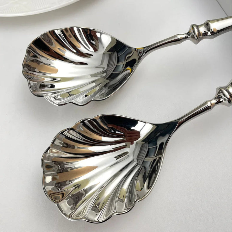 A pair of ornate, polished silver serving spoons with scalloped, shell-shaped heads.