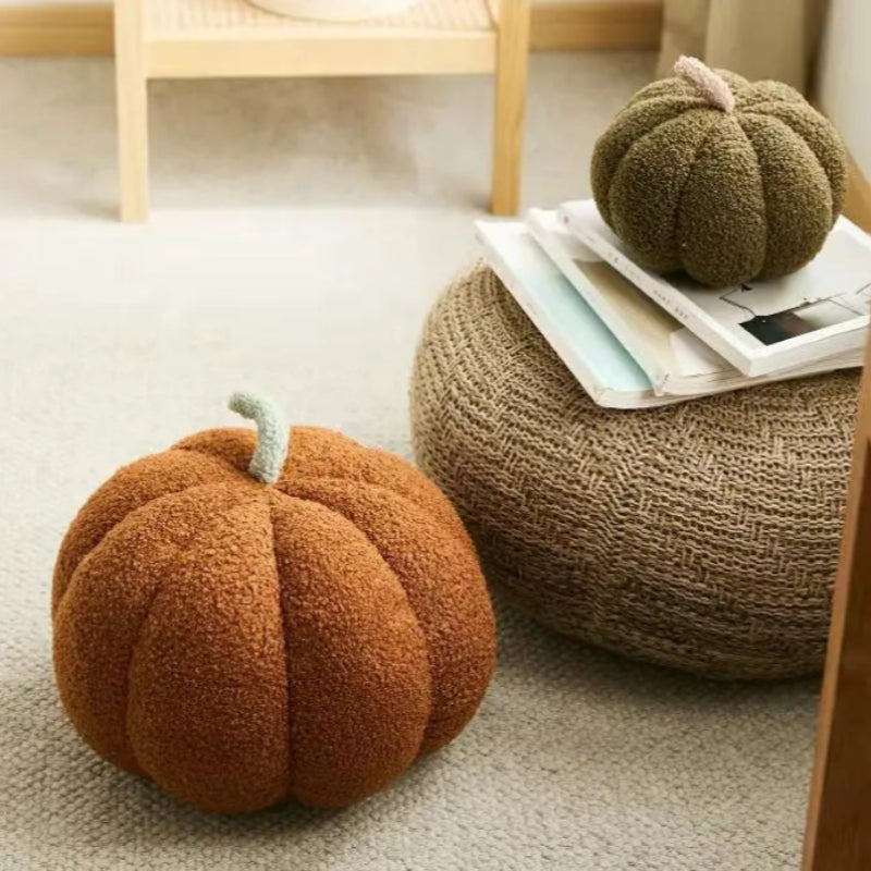 Two soft, pumpkin-shaped pillows, one brown on the floor and one green atop a stack of books on a woven stool, are featured in a cozy indoor setting.