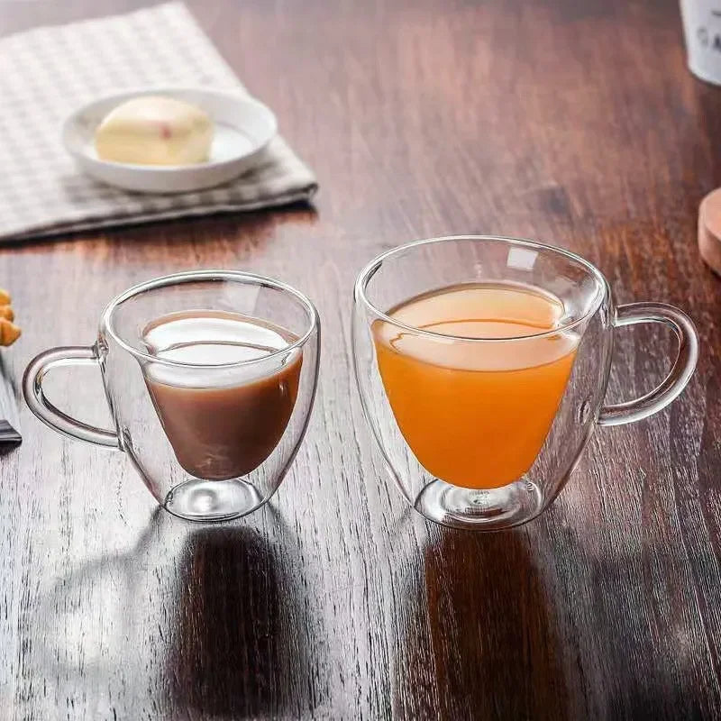 Two heart-shaped glass mugs on a wooden table, one with coffee and the other with orange juice, with a plate of pastries in the background.