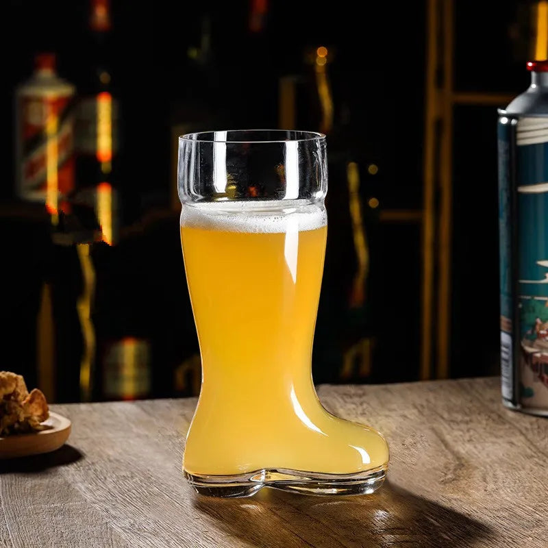 A clear glass beer boot mug filled with a light-colored beer and white foam sits on a wooden surface in a dimly lit bar setting with blurred bottles in the background.