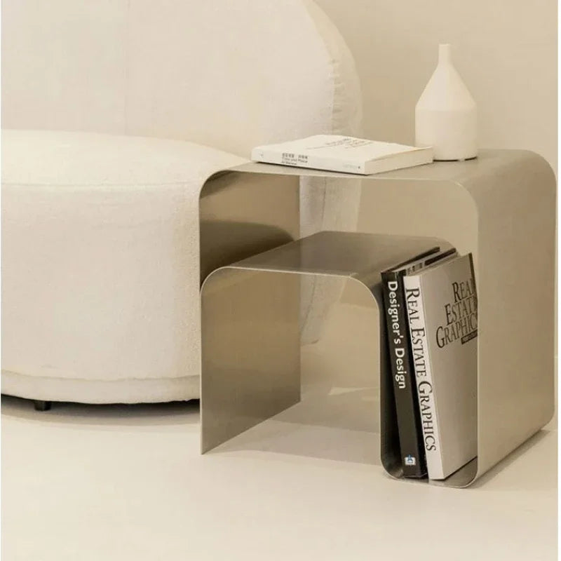A shiny, metallic silver side table with rounded corners holds a smaller table inside it, with books tucked between them. A white couch and vase are in the background.
