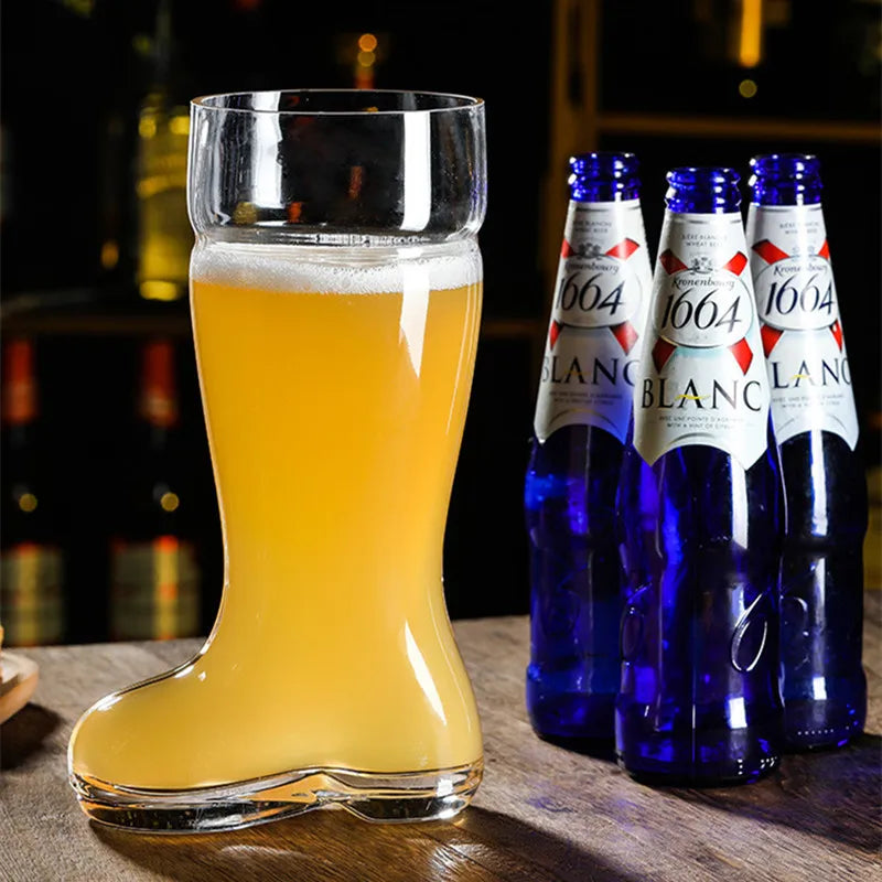 A clear glass beer boot mug filled with light-colored beer and white foam sits on a dark surface next to four blue glass beer bottles with white labels in a dimly lit setting.
