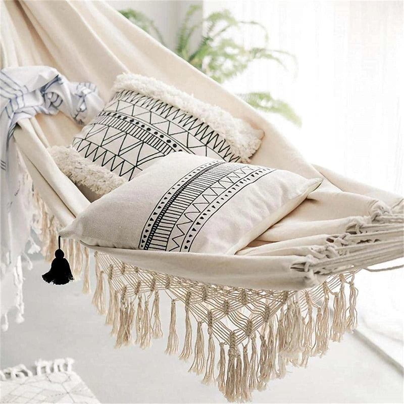 A beige fabric hammock with macrame fringe and a black tassel is adorned with two pillows featuring black geometric patterns and a striped throw blanket.