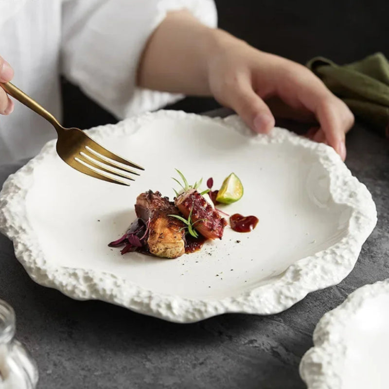 A person holding a gold fork over a white, textured plate with a beautifully plated dish of seared meat and a lime wedge.