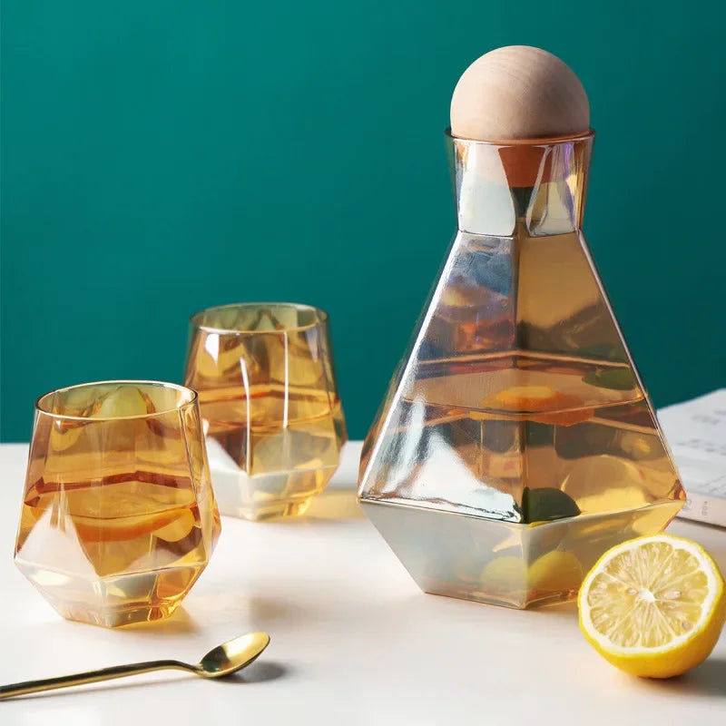 Amber-tinted glass pitcher and two matching faceted glasses on a white surface, with a gold spoon and a lemon slice. The background is a dark teal color.