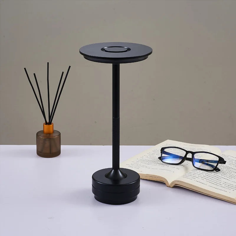 A sleek black desk lamp with a circular flat top and a textured handle, standing on a white table. An open book with black-framed glasses rests beside it, while a reed diffuser in a brown glass container is positioned in the background.