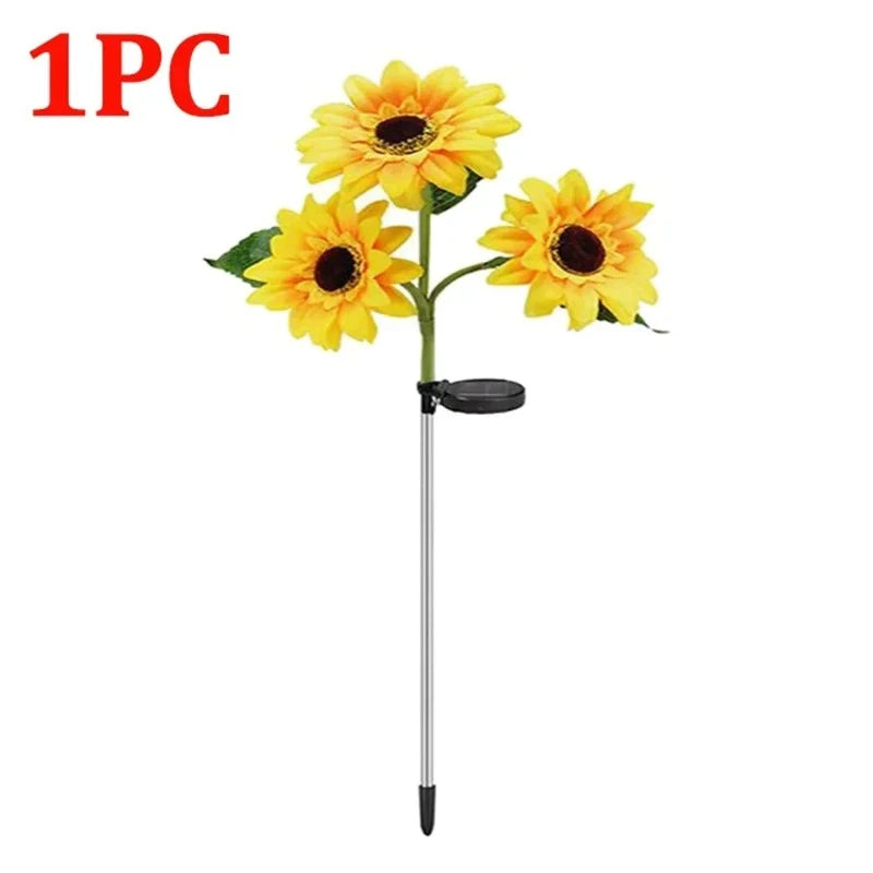 Three artificial sunflowers with bright yellow petals and dark brown centers bloom on a green stem with a few green leaves. A black solar panel is attached to the stem. The base of the stem is a silver stake with a pointed black end. "1PC" is visible in the upper left corner against a white background.