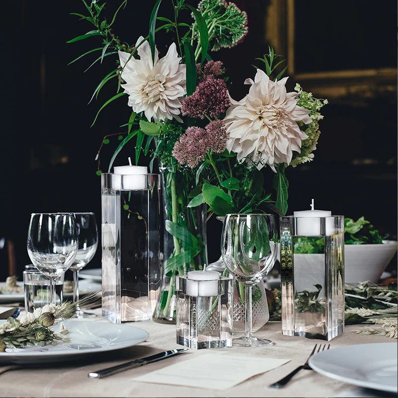 A formally set table features a large clear glass vase filled with various white and burgundy flowers and green foliage as a centerpiece. Clear, rectangular candle holders with white pillar candles are placed around the vase. Wine glasses and water glasses are set at each place setting, along with white plates, silverware, and light-colored napkins. The background is dimly lit, suggesting an elegant dining atmosphere.