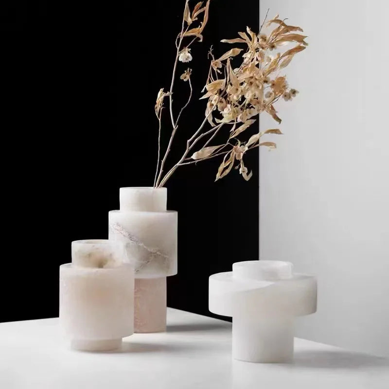 A collection of Scandinavian White Onyx vases displayed on a white surface against a contrasting black-and-white background. The vases feature geometric shapes and natural marble veining, with one elegantly holding dried floral branches, adding an artistic touch to the modern arrangement.