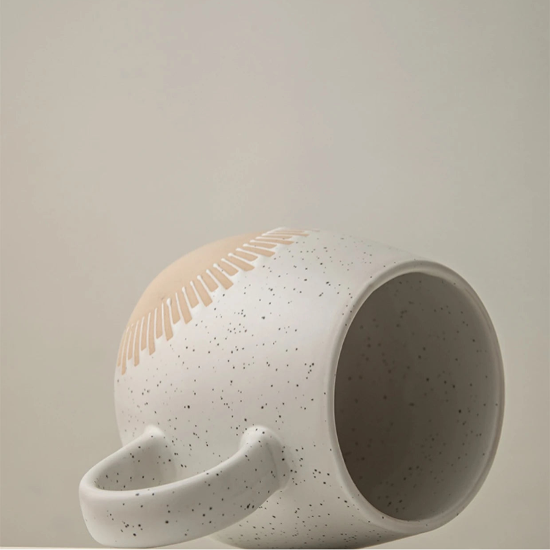 A speckled ceramic mug with an embossed sunburst design is lying on its side on a light-colored surface. The mug has a curved handle and a smooth, minimalist finish with a modern aesthetic.