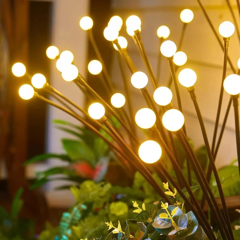 Several slender dark stems rise from vibrant green foliage, each culminating in a glowing warm white spherical light. The lights are positioned at varying heights, creating a whimsical, firefly-like effect in a garden setting at dusk or dawn.