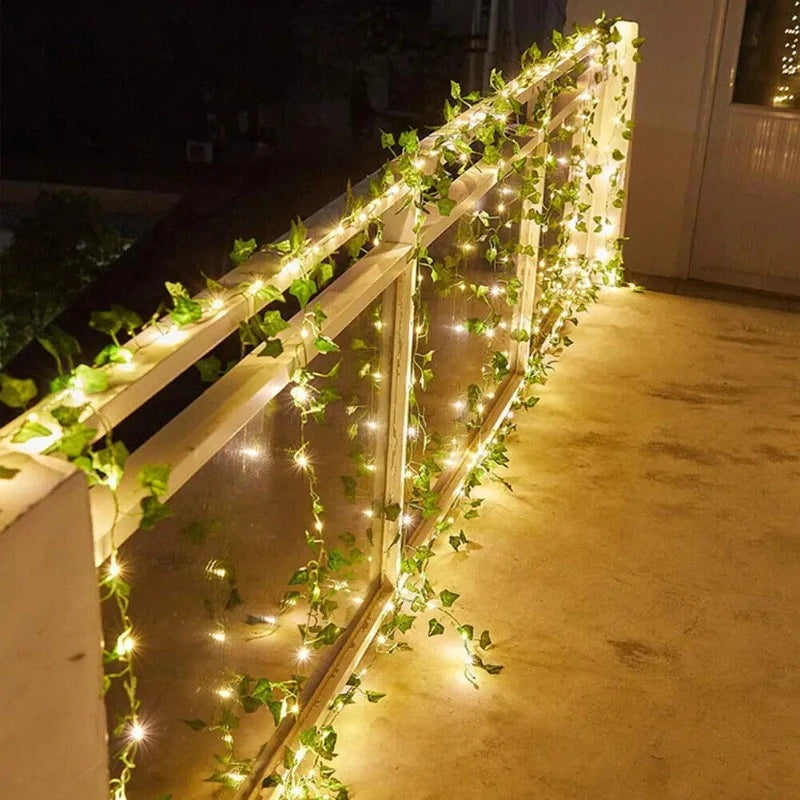 A string of warm white solar-powered LED lights intertwined with artificial green ivy leaves is wrapped around a white outdoor railing at night. The lights illuminate the railing and the surface below.